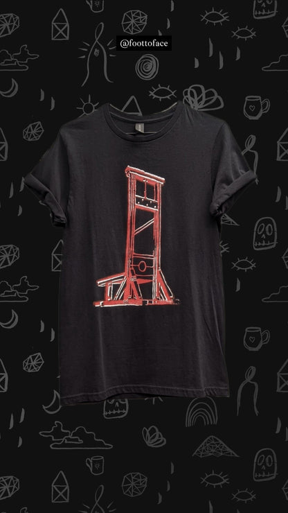 A black short-sleeve t-shirt featuring a red hand-drawn sketch of a guillotine, displayed against a dark background patterned with white doodles and the brand name @foottoface.
