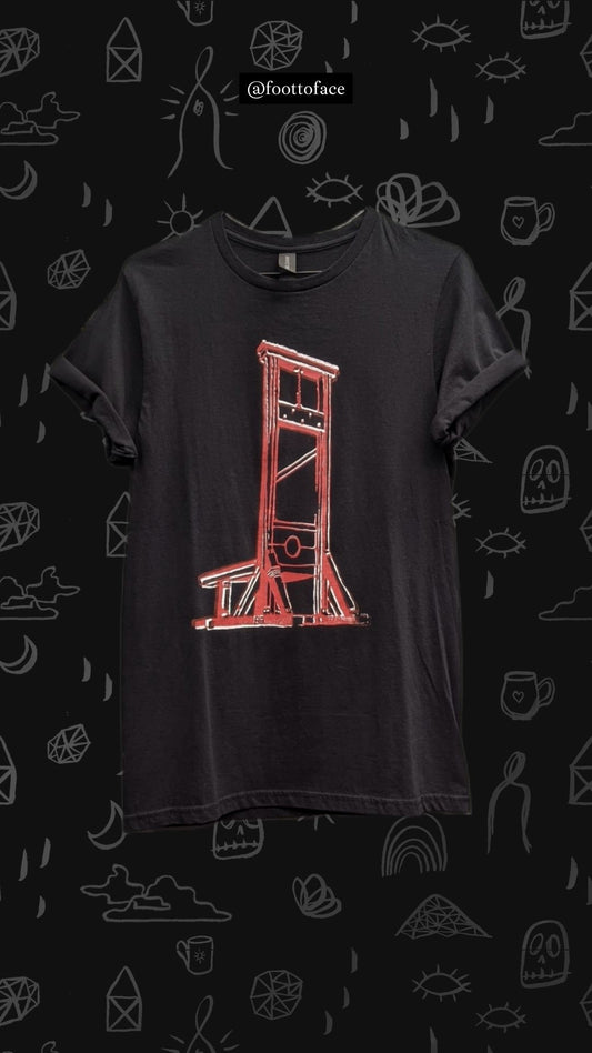 A black short-sleeve t-shirt featuring a red hand-drawn sketch of a guillotine, displayed against a dark background patterned with white doodles and the brand name @foottoface.